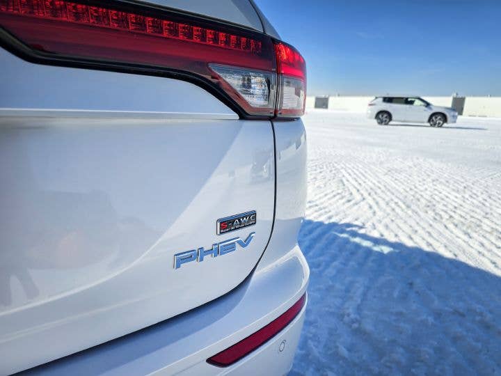 A closeup of the Outlander PHEV badge with a white Outlander PHEV in the distance